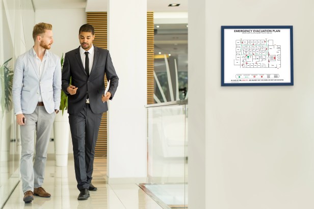 A facility manager stands in a clean, modern office building lobby looking at a clipboard with a checklist. A Bordered Evacuation Map is clearly visible on the wall. Conveys proactive safety and effective management.