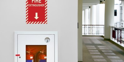 A modern office hallway with a bright red Fire Extinguisher w/ Arrow - LaserGlow (EMS-FE75-D) mounted on the wall. A "FIRE EXTINGUISHER" sign with a directional arrow is positioned directly above it.
