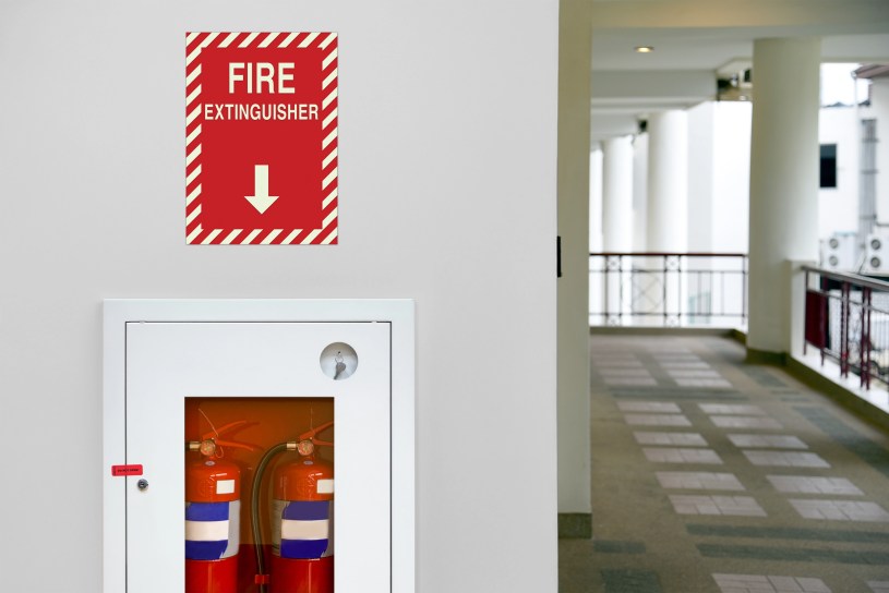 A modern office hallway with a bright red Fire Extinguisher w/ Arrow - LaserGlow (EMS-FE75-D) mounted on the wall. A "FIRE EXTINGUISHER" sign with a directional arrow is positioned directly above it.