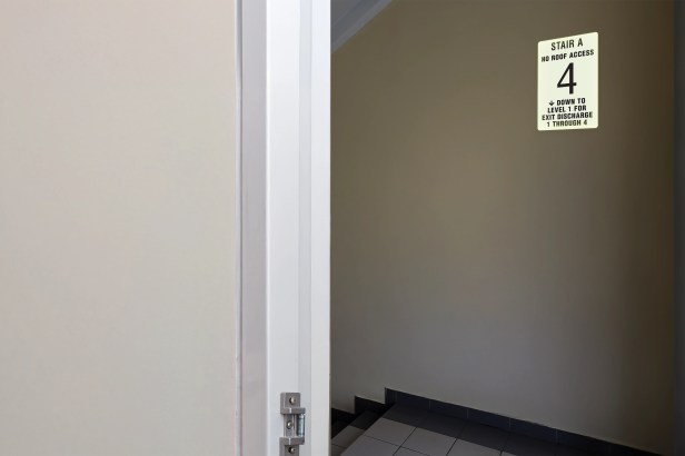 A Photoluminescent Stairwell ID • 4 Floors or More (NFPA-PVC1812) sign glows faintly in a dark stairwell, demonstrating visibility during a power outage. The background is blurred, focusing attention on the sign itself.