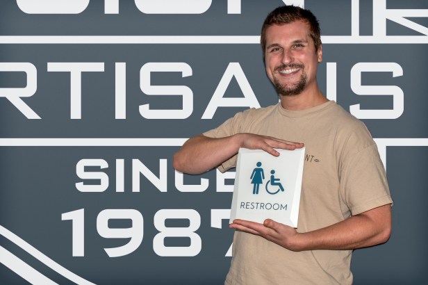 EvacuationPlans.com artisan Hunter Higgins presenting a Facets ADA-compliant restroom sign, featuring precision-cut angles and tactile lettering designed for accessibility and wayfinding compliance.