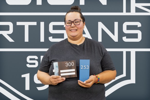 EvacuationPlans.com artisan Michelle Garrett displaying two interior ADA-compliant signs from the Botanica and Aleigha lines, demonstrating refined design and clear readability for safe building navigation.