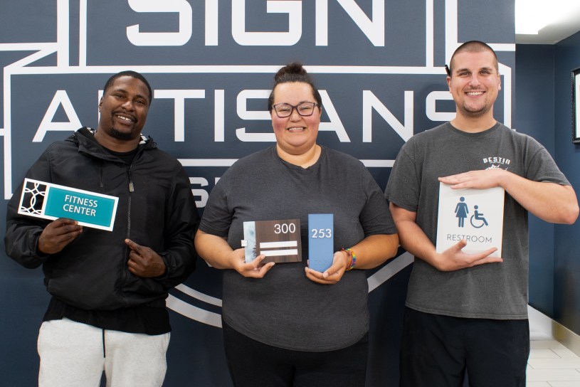EvacuationPlans.com artisans Eric Reynolds, Michelle Garrett, and Hunter Higgins holding custom ADA-compliant signs, representing the teamwork and craftsmanship behind safe, code-ready building signage.