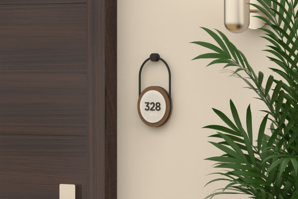 A 3D-printed RN95 room number sign featuring raised “328” numerals and braille, displayed on a rounded plaque suspended from a matte black hanging mount. The sign combines a White (S02) face with Spanish Leather (S17) sides and Black (S37) tactile lettering, creating a warm, modern hospitality aesthetic beside a dark wood door and indoor greenery.
