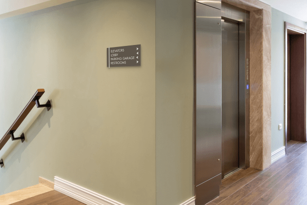A Midtown 4-Line Directional Sign (MID-D611) mounted near an elevator in a multi-story facility, listing Elevators, Lobby, Parking Garage, and Restrooms with directional arrows. The sign features an Antique Bronze (S96) faceplate with Oyster (S90) accents, providing clear, code-aligned wayfinding to support safe movement during daily operations and emergency events.