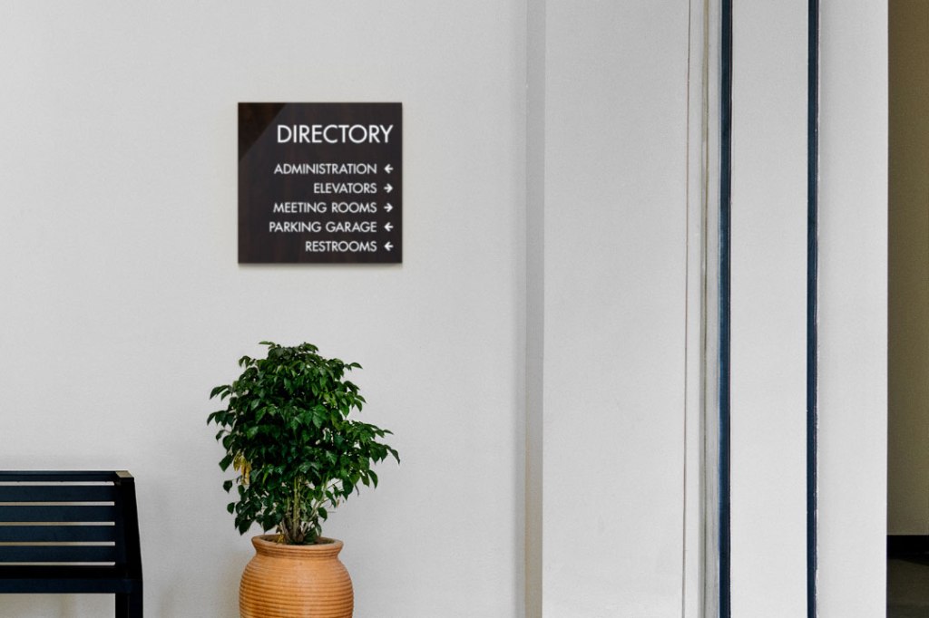 Wall-mounted directory sign showing directions for administration, elevators, meeting rooms, parking garage, and restrooms.