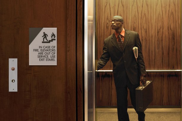 A man stands inside an elevator holding a briefcase while a wall-mounted ‘In Case of Fire, Elevators Are Out of Service—Use Exit Stairs’ sign is displayed beside the elevator doors.