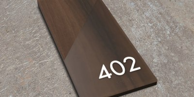 A modern ADA-compliant room number sign in the Incline Sign Style, featuring a diagonal two-tone wood design with raised tactile numbers and Grade 2 braille reading ‘402,’ displayed on a neutral textured surface.