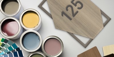 A flat lay of paint cans, color swatches, and woodgrain samples arranged around a modern room number plaque reading “125.” The image highlights customization options, material finishes, and color variety used in creating professional interior signage.