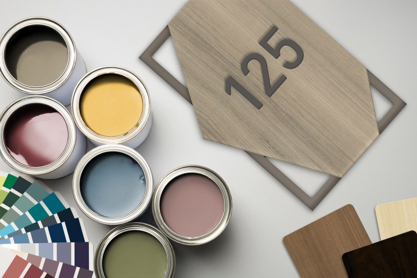 A flat lay of paint cans, color swatches, and woodgrain samples arranged around a modern room number plaque reading “125.” The image highlights customization options, material finishes, and color variety used in creating professional interior signage.