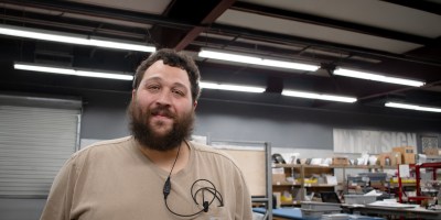 Nick Cruz, EvacuationPlans.com Sign Artisan of the Month, standing in the EvacuationPlans.com production facility surrounded by worktables and materials, representing the craftsmanship behind custom signage.