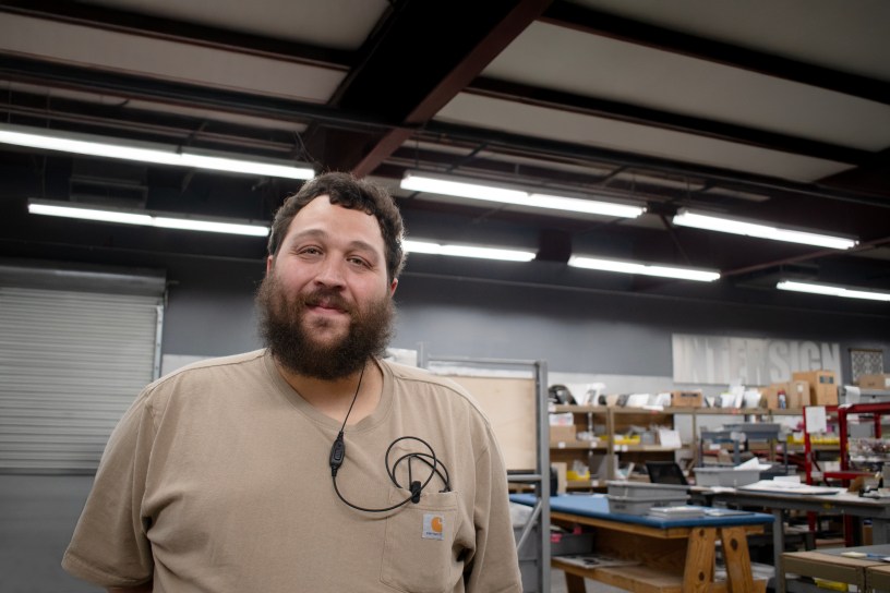 Nick Cruz, EvacuationPlans.com Sign Artisan of the Month, standing in the EvacuationPlans.com production facility surrounded by worktables and materials, representing the craftsmanship behind custom signage.